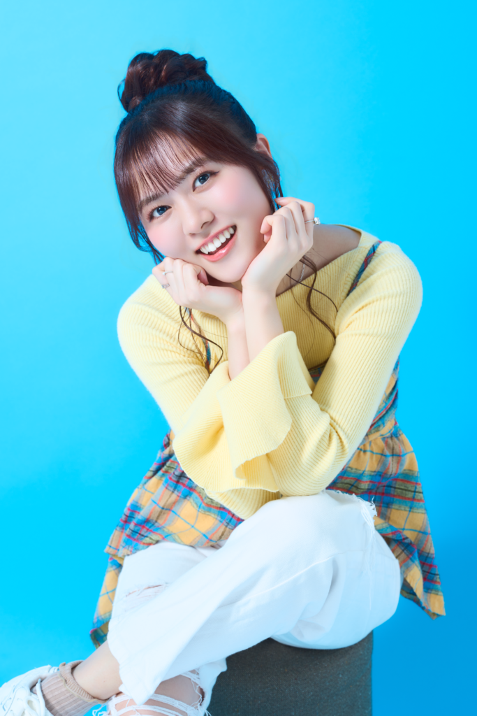 Cheerful young woman with dark hair in a bun, wearing a yellow knit sweater and plaid skirt, posing with hands under her chin against a bright blue background.