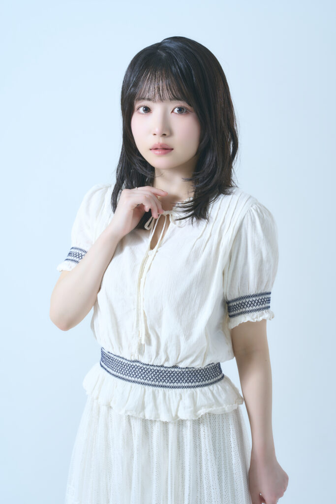 Portrait of a young woman with shoulder-length dark hair, wearing a white textured dress, looking at the camera with a soft expression against a pale blue background.