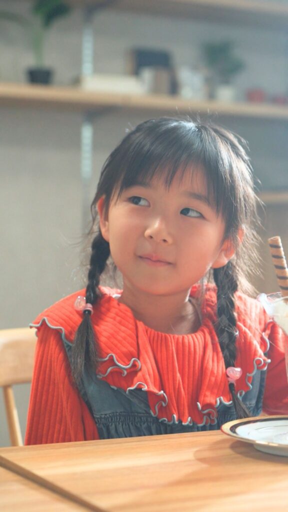 Three-quarter view of a young girl with braided pigtails wearing a red knit top and denim overalls, sitting at a wooden table indoors and looking to the side.