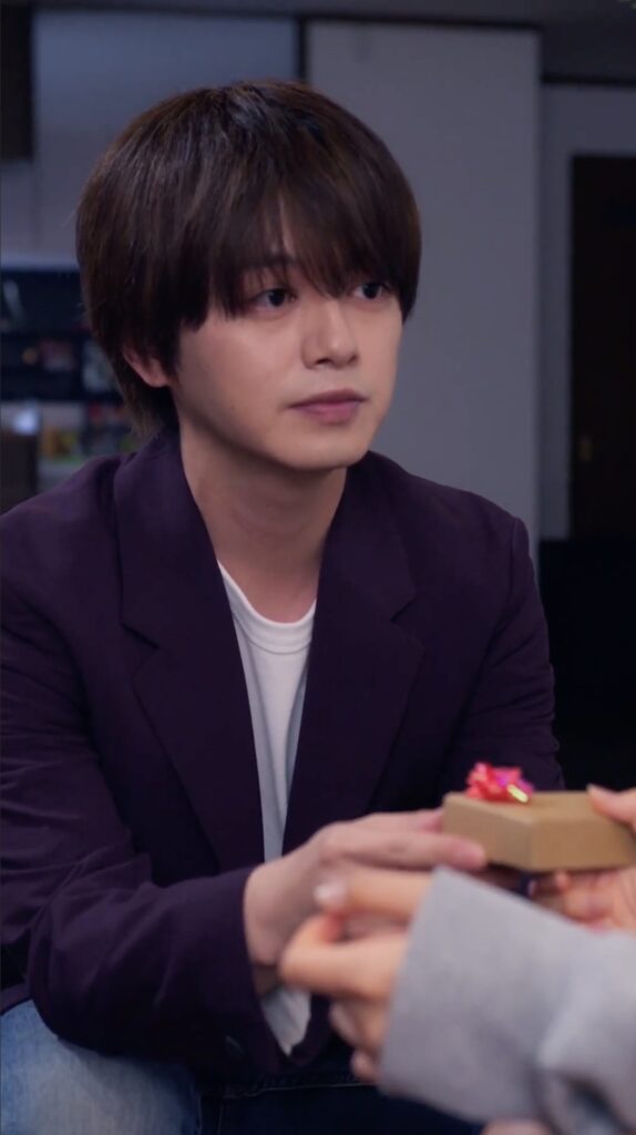 Young man with dark hair wearing a purple blazer sits and receives a small gift box with a red bow.