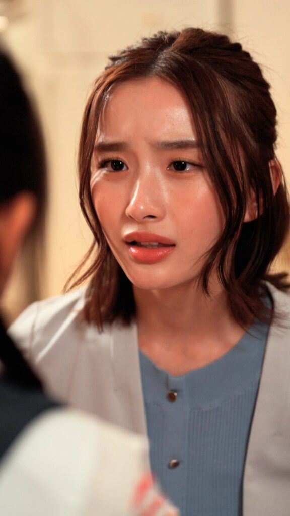 Close-up of a young woman with brown hair speaking to someone off-camera, wearing a blue top and light cardigan.