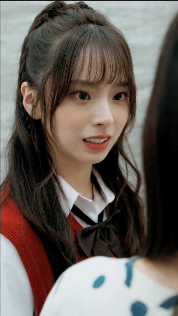 Close-up of a young woman with long dark hair in a red sweater vest over a white blouse, wearing a black bow, looking to the side.