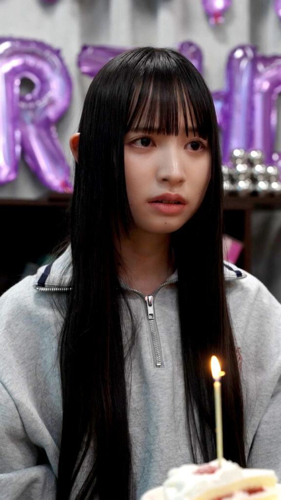 Young woman with long black hair in a gray zip-up hoodie sits at a birthday cake with a lit candle, purple balloons in the background.