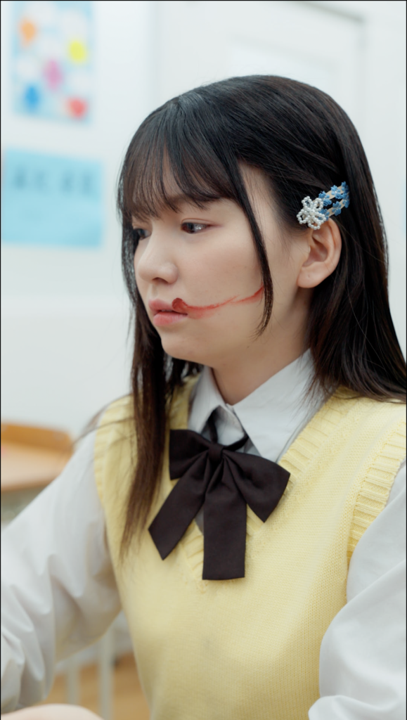 Young girl in a yellow knit vest over a white shirt with a dark bow, looking to the left, with red face makeup forming a scar across her mouth.