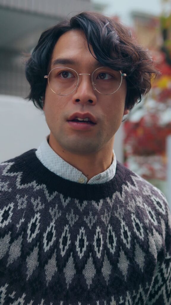 Young man with wavy dark hair and round glasses, wearing a dotted white shirt under a black-and-gray patterned sweater, looking to the side outdoors.