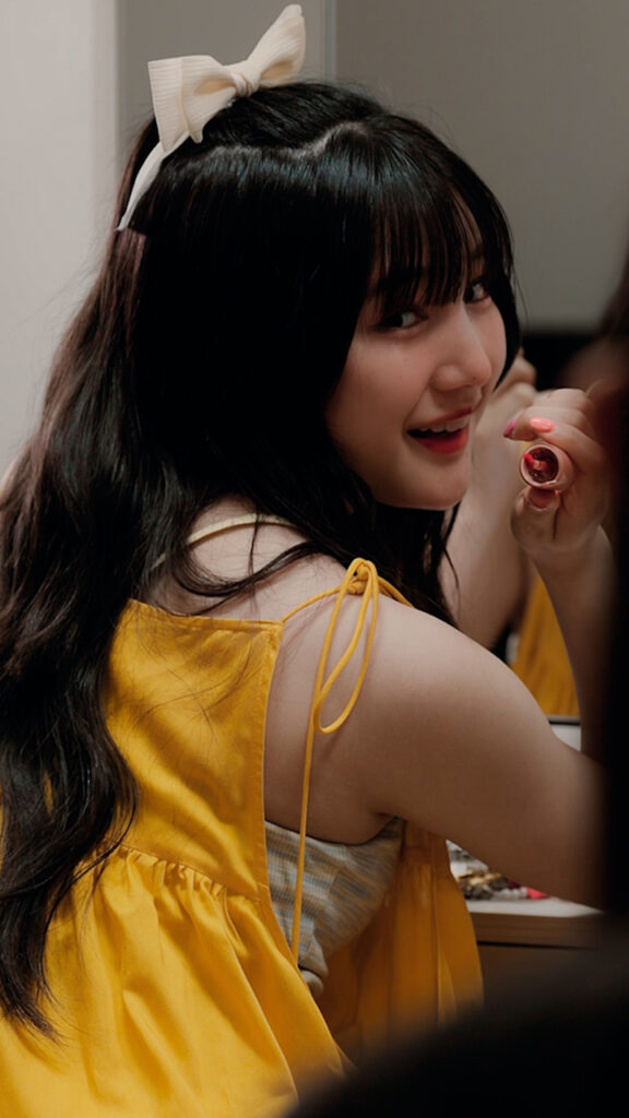 Young woman with long black hair and a white bow in a yellow dress, smiling while applying lipstick in a mirror-like setting.