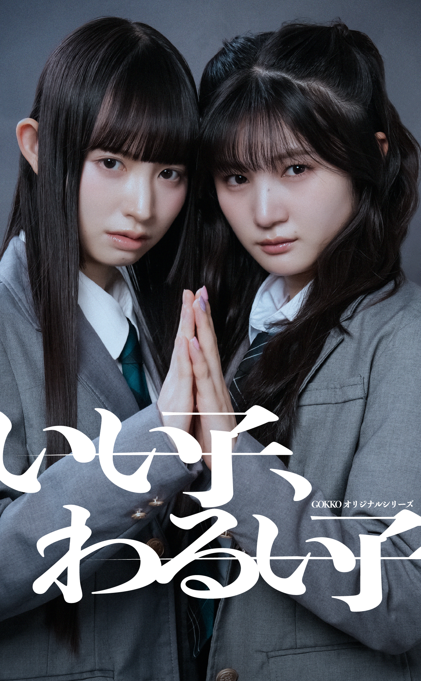 Two young women in gray school uniforms press their hands together and look at the camera against a dark backdrop, posing for a promotional image.