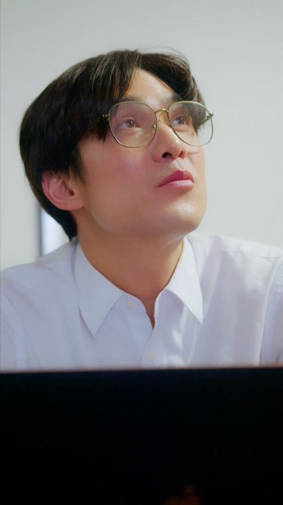 Sc - 縦型ショートドラマなら GOKKO Inc.|ごっこ倶楽部 Close-up of a man wearing round glasses and a white shirt, looking upward with a thoughtful expression in an office setting.