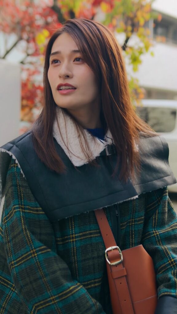 Portrait of a woman with shoulder-length brown hair, wearing a plaid coat with a shearling collar, outdoors among autumn leaves.