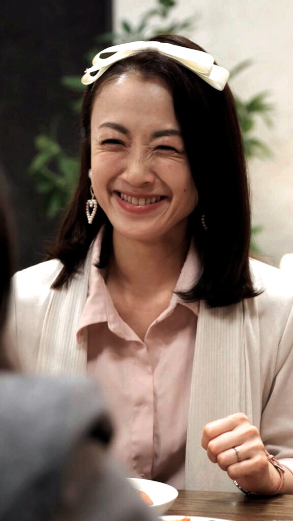 Woman smiling at a table, wearing a white headband, pink blouse, and cream cardigan; a bowl is in the foreground.