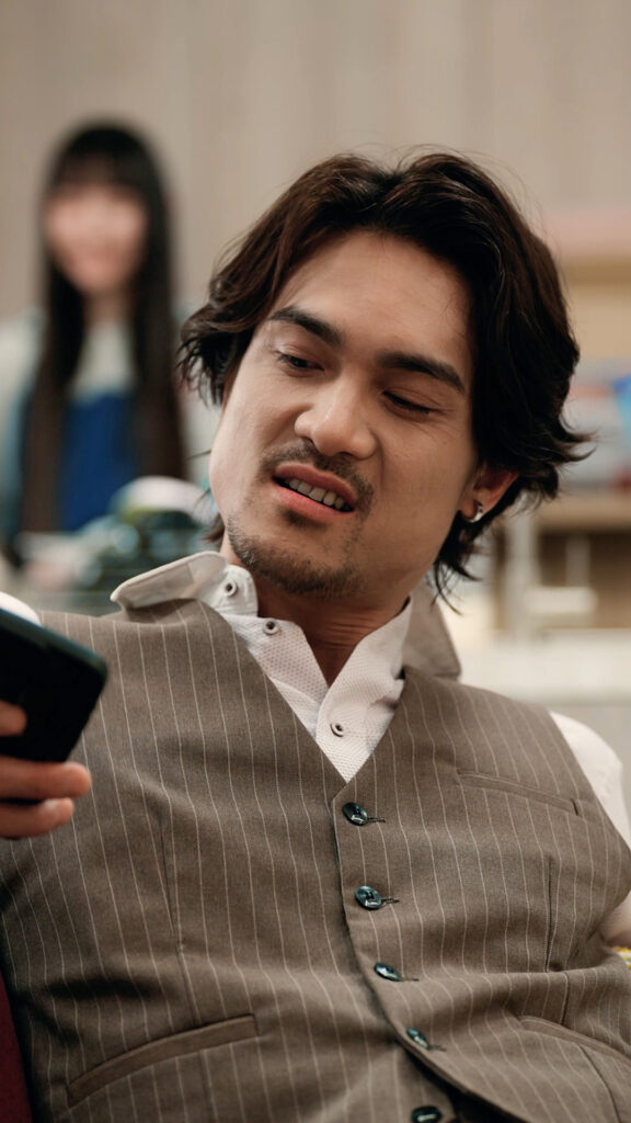 Man in a brown pinstripe vest looks at a smartphone indoors; a blurred woman stands in the background.