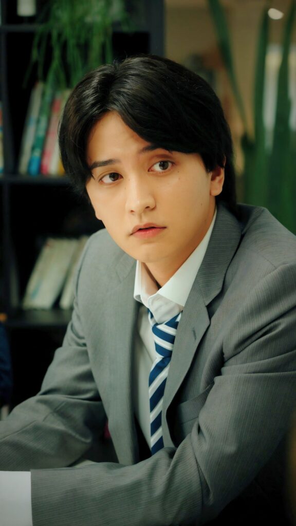 瀬戸利樹 様_E - 縦型ショートドラマなら GOKKO Inc.|ごっこ倶楽部 Portrait of a young man in a grey suit and striped blue tie, seated at a desk in a library or office, looking off to the side.