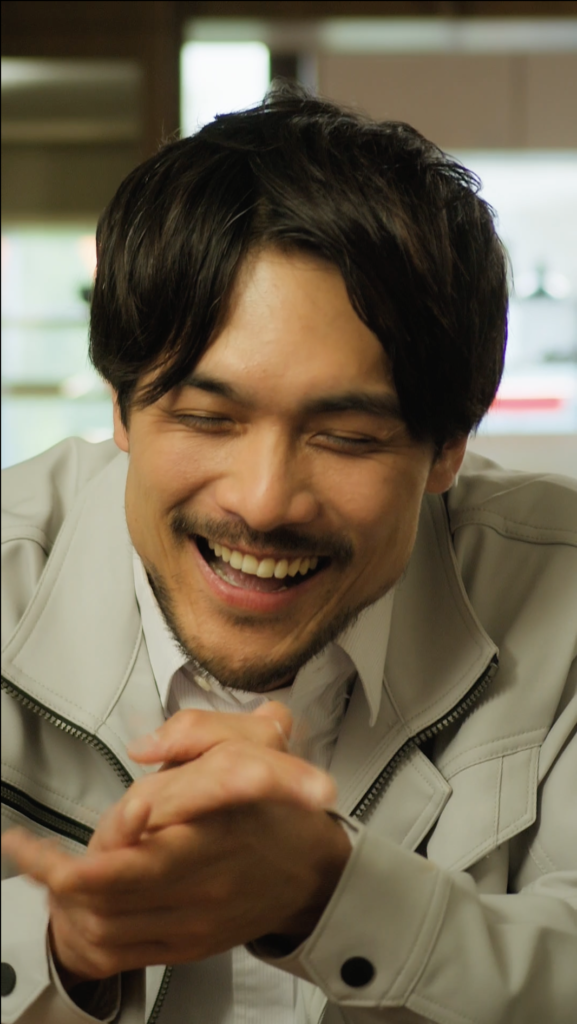 谷沢龍馬さま04 - 縦型ショートドラマなら GOKKO Inc.|ごっこ倶楽部 Close-up of a man with dark hair and stubble laughing, hands clasped together in front, wearing a light jacket.