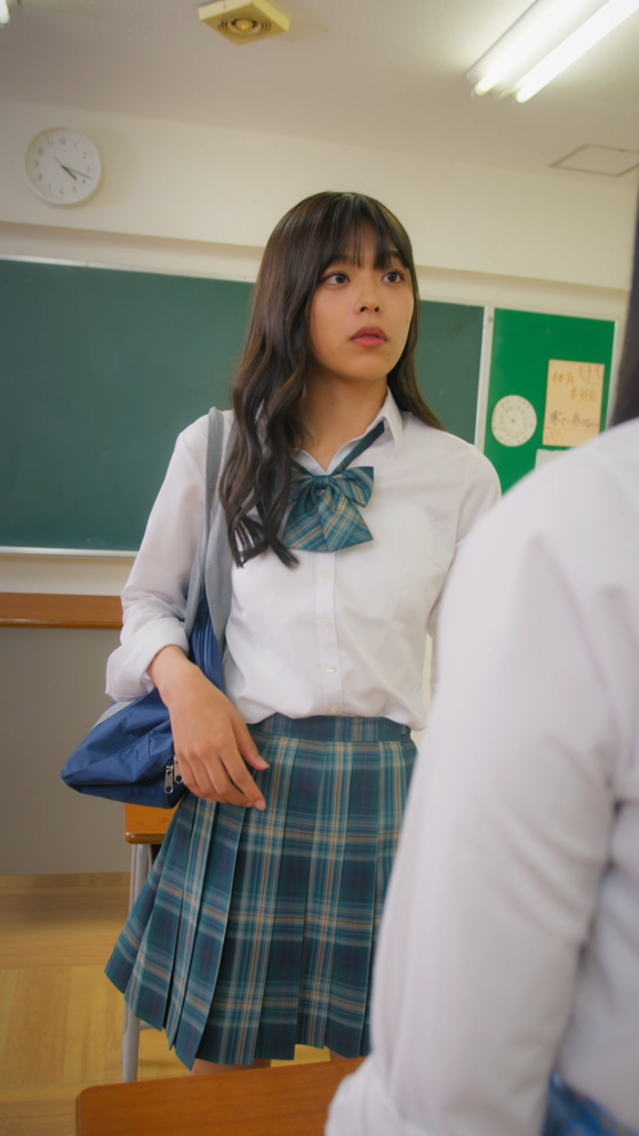 川田明日未さま3 - 縦型ショートドラマなら GOKKO Inc.|ごっこ倶楽部 Schoolgirl in a white blouse and teal plaid skirt standing by a desk in a classroom, carrying a blue bag.