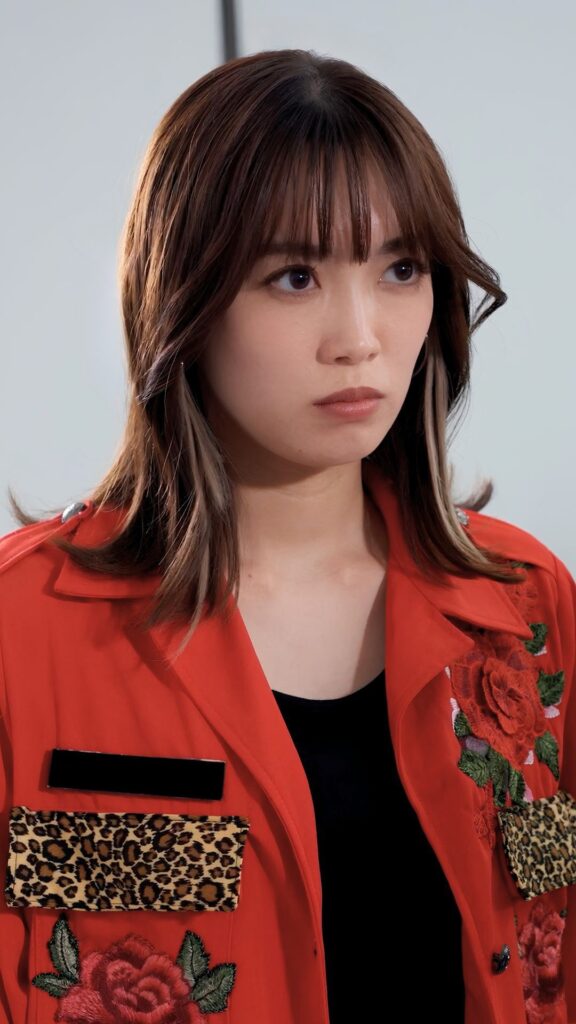 Woman with shoulder-length brown hair and bangs wearing a red embroidered jacket, looking to the side.