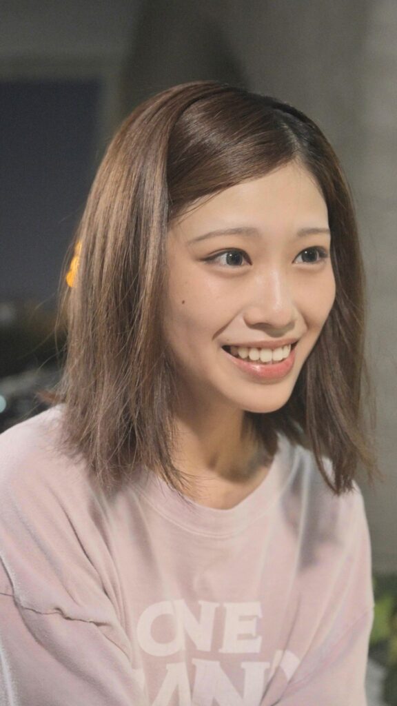 中里芙紅 様_C - 縦型ショートドラマなら GOKKO Inc.|ごっこ倶楽部 Young woman with shoulder-length brown hair smiling softly, wearing a pink sweatshirt, in a blurred indoor setting.