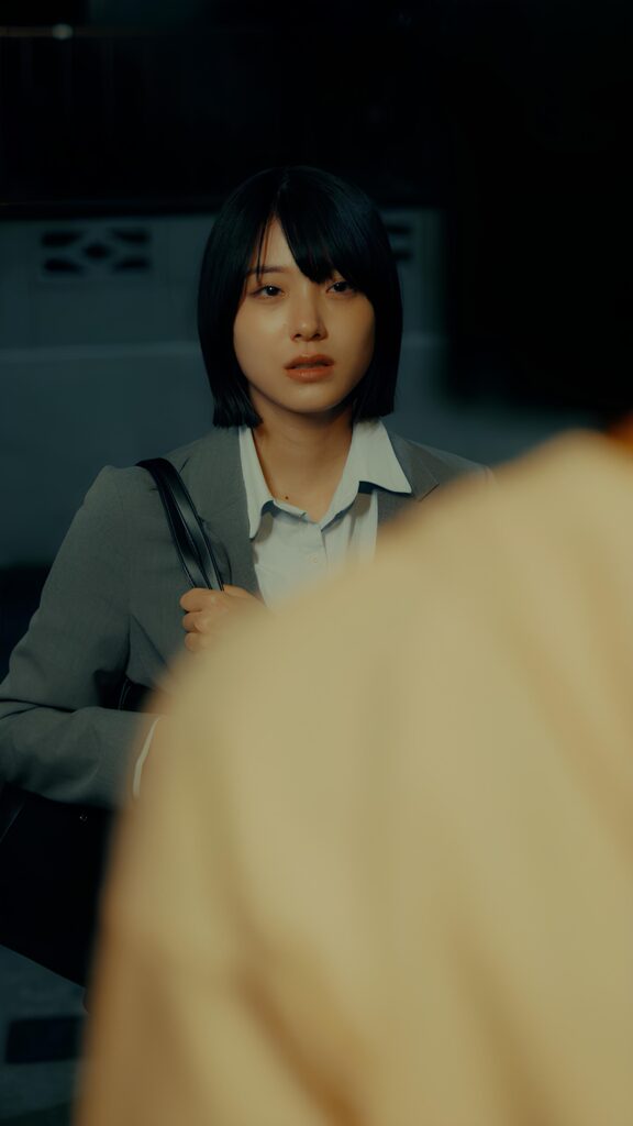 岩波詩織様_A - 縦型ショートドラマなら GOKKO Inc.|ごっこ倶楽部 Young Asian woman in a gray blazer and white shirt, carrying a black bag, in a dim indoor setting.