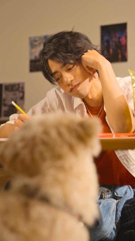 後藤光輝 様_E - 縦型ショートドラマなら GOKKO Inc.|ごっこ倶楽部 Student with dark hair leaning on his hand while writing with a pencil at a desk in a classroom.