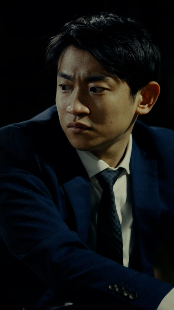 早坂架威様_I - 縦型ショートドラマなら GOKKO Inc.|ごっこ倶楽部 Asian man in a navy blazer and tie, looking to his left with a serious expression against a dark background.