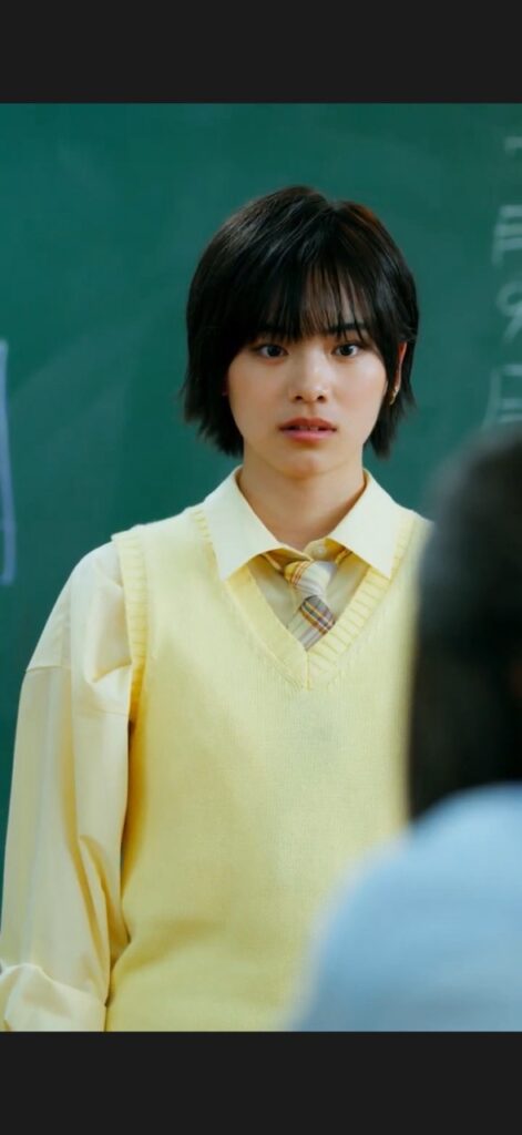 Student with short dark hair wearing a yellow sweater vest and collared shirt stands in a classroom with a chalkboard behind them