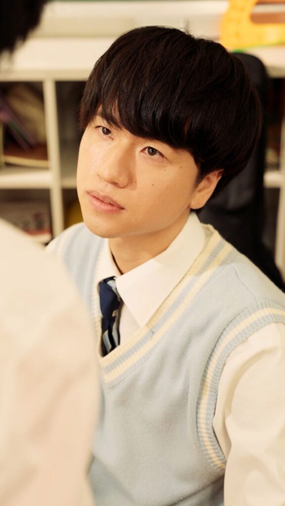根井深考 様_C - 縦型ショートドラマなら GOKKO Inc.|ごっこ倶楽部 Young man with dark hair wearing a light blue sweater vest over a white shirt and navy tie, looking to the side indoors.