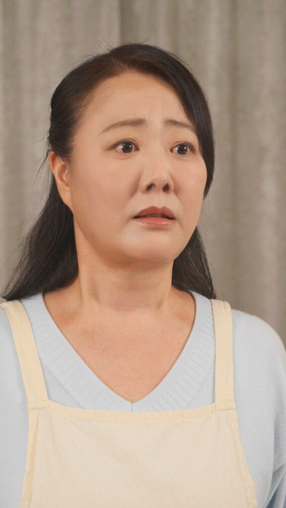 色味修正済水月舞 様_C - 縦型ショートドラマなら GOKKO Inc.|ごっこ倶楽部 Asian woman with worried expression, wearing a light blue top and beige apron, looking off to the side against a neutral background.