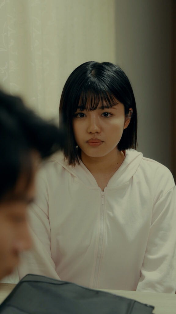 舞花様_L - 縦型ショートドラマなら GOKKO Inc.|ごっこ倶楽部 Young woman with short black hair in a light hoodie sits at a table, looking downward, with a blurred person in the foreground.